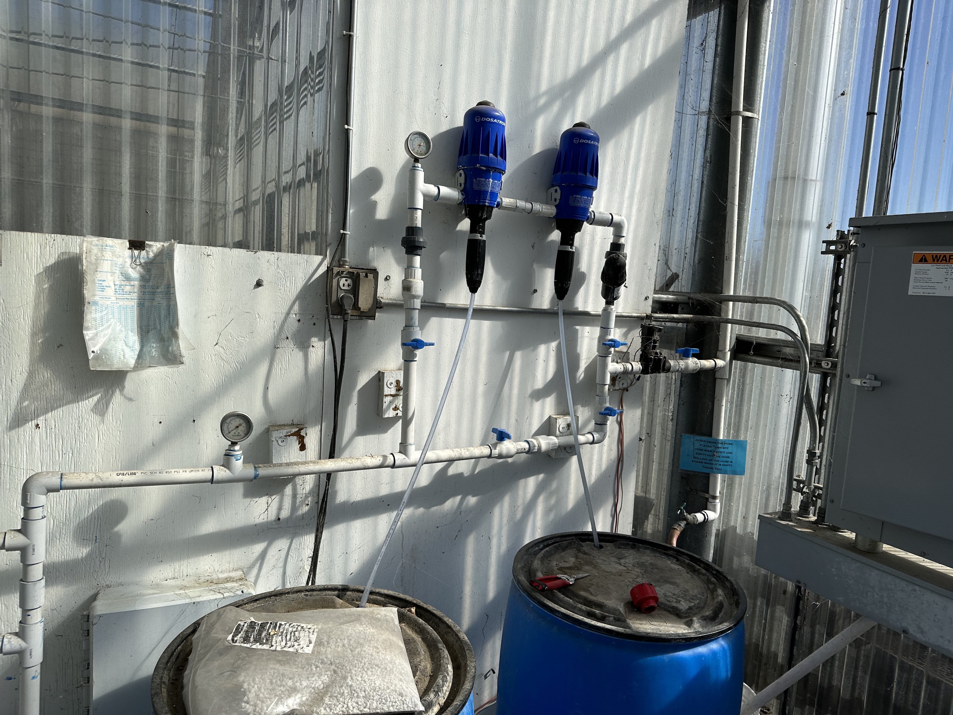 Two fertilizer proportioners mounted on a wall connected to the irrigation system and two barrels with pvc pipe.