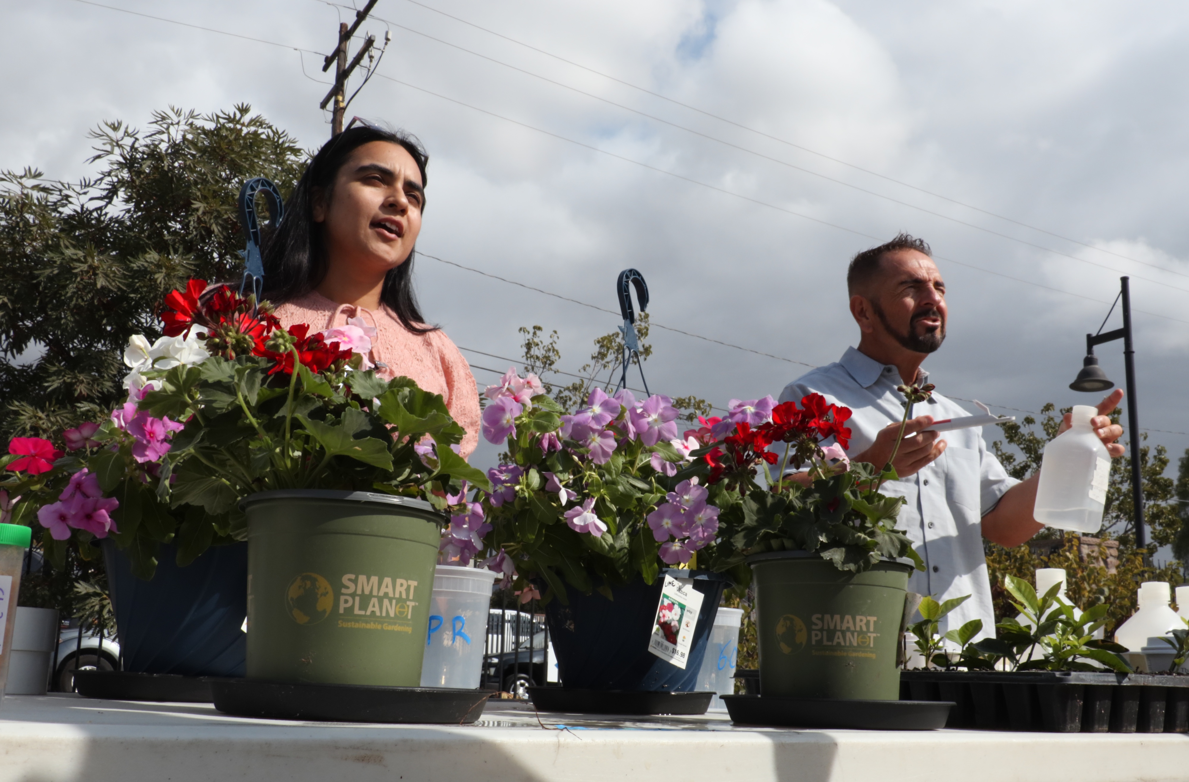 A photo of two presenters standing behind a table with potted flowers.