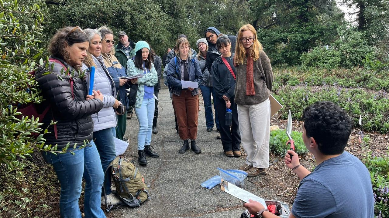 A photo of Dr. Sebastian Fajardo kneeling in a garden holding a clipboard and trowel in front of a group of workshop participants.