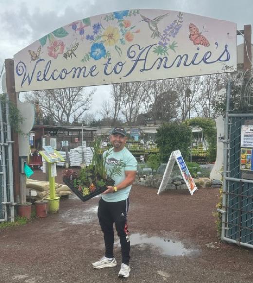 A photo of Don Merhaut smiling and holding a tray of healthy plants under an arching sign into a nursery that says Welcome to Annie's in stylized script with painted flowers, birds, and butterflies