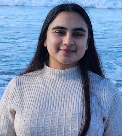 Profile Photo of Haramrit Gill. A woman with long black hair wearing a white sweater standing in front of a body of water.