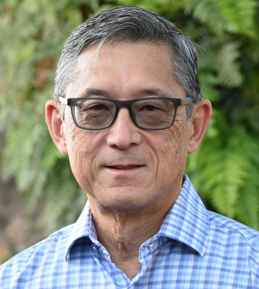 Profile Photo of Lorence Oki. A man with grey hair and brown glasses wearing a blue checkered button up shirt.